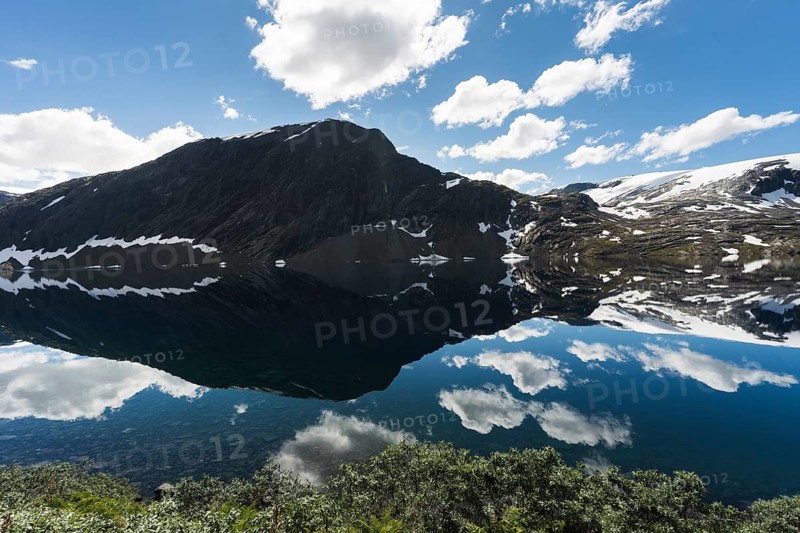 A serene mountain lake in Norway mirrors the dramatic peaks, snow patches, and a bright sky The tranquil waters and natural beauty make it a perfect scene for adventure and peace