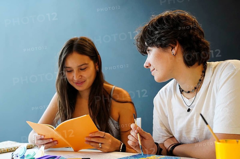 A non-binary teenager with short hair and a female teenager engage in a study session, displaying concentration and friendship. They are both focused on educational materials at a table, surrounded by art supplies