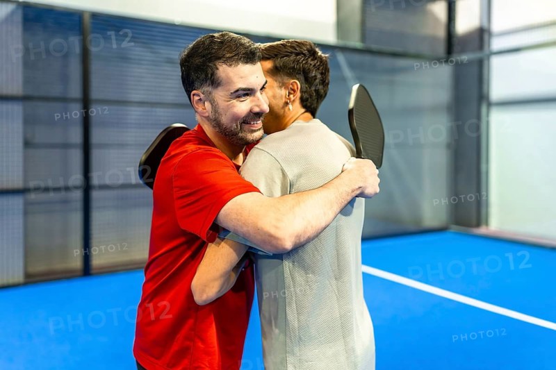 Two adult men, wearing sportswear and holding paddles, embracing on a bright blue pickleball court, showing friendship, camaraderie, and celebrating a game