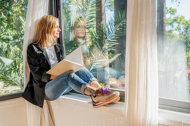 A woman sits by the window, reading a book, with lush greenery outside She enjoys a serene atmosphere, wearing casual jeans and a blazer, reflecting relaxation and focus