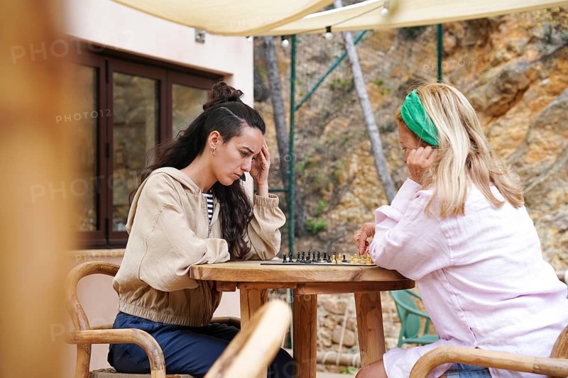Mother and daughter are deeply focused on a chess game outdoors, surrounded by nature They sit at a wooden table under shade, enjoying a peaceful sunny day
