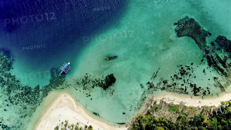 An aerial view captures two boats floating on the clear, turquoise waters near a white sandy beach in the Philippines, surrounded by lush greenery and coral reefs