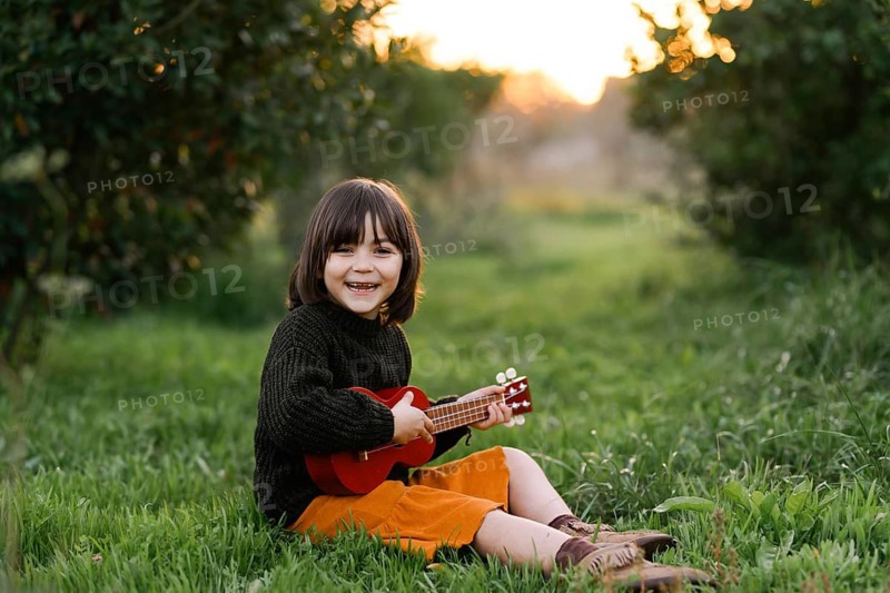 A cheerful child sits in a grassy meadow, strumming a small red guitar. The vibrant scene captures joy and creativity in a natural setting, surrounded by lush greenery