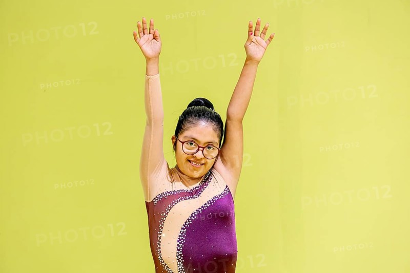 A young disabled gymnast stands confidently with arms raised against a bright green backdrop. She wears glasses and a purple leotard, embodying enthusiasm and determination