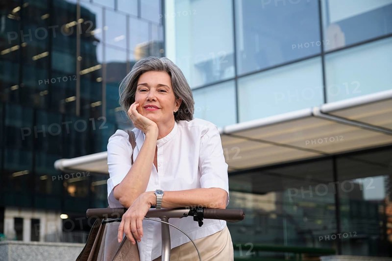 A confident mature woman with gray hair leans on an electric scooter, smiling warmly. She is outside a modern glass building, enjoying a peaceful, sunny day in the city