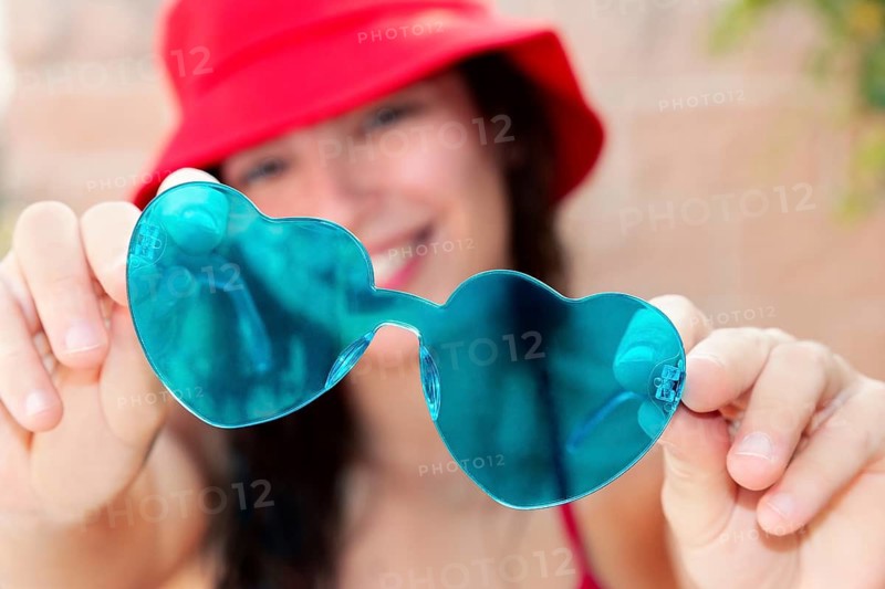 A Generation Z girl holds up heart shaped sunglasses, smiling in the bright summer sun. Her vibrant red hat and cheerful demeanor capture the youthful spirit of the season