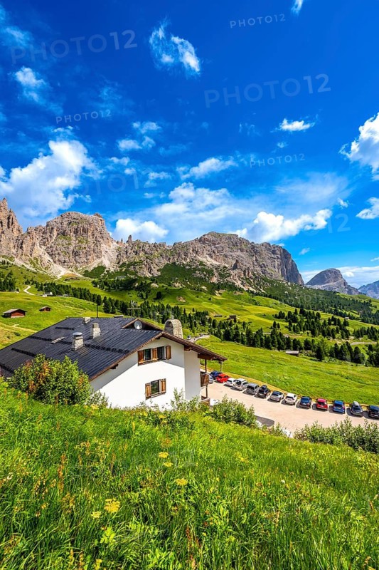 Scenic view of a mountain chalet with parked cars, surrounded by lush greenery and the majestic peaks of the dolomites in summer