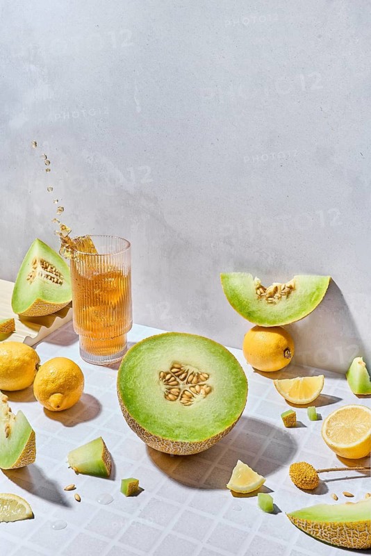 Fresh melon and lemon slices and fresh juice in a glass arranged creatively on a light, textured surface. The soft sunlight and vibrant colors evoke a refreshing summer vibe and healthy lifestyle feel