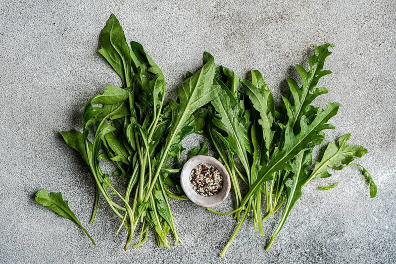 Fresh arugula leaves spread on a textured concrete surface with a small bowl filled with mixed seasoning. The vibrant green leaves contrast with the rustic background