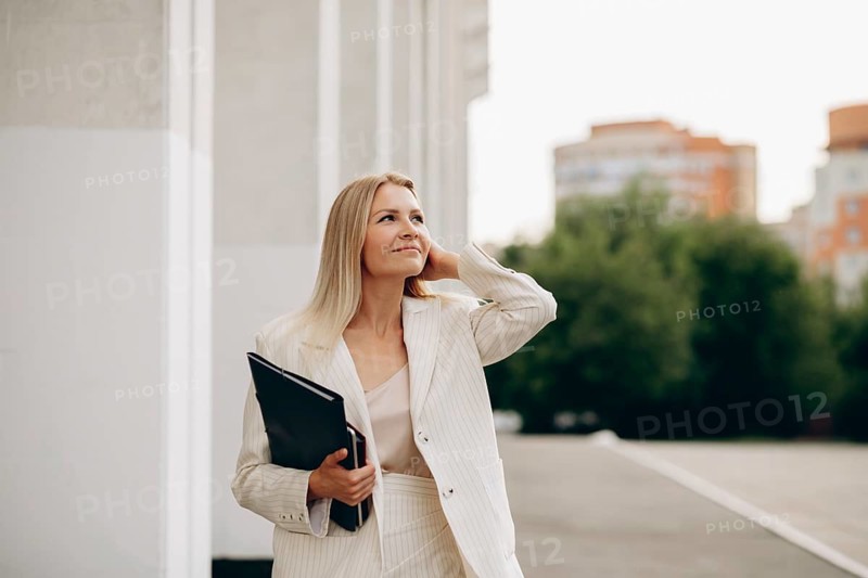 A confident businesswoman in a white suit holds folder while walking in an urban area. The setting features modern architecture and lush greenery under a bright sky