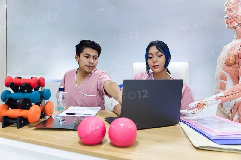 Two physiotherapists in a modern Ecuadorian clinic discuss treatment plans over a laptop, with anatomy models and exercise equipment nearby, signifying a collaborative healthcare approach