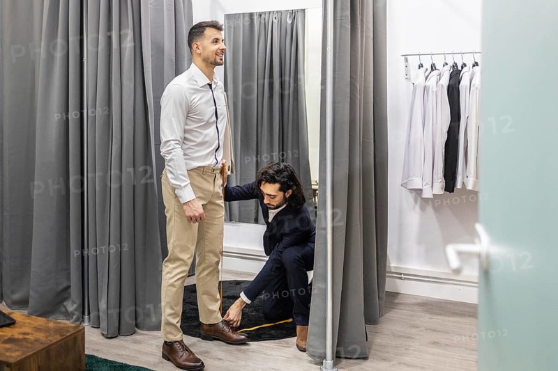 A professional tailor measures a man for pant fitting in a stylish fitting room. The customer stands smiling in front of a mirror, surrounded by shirts on hangers
