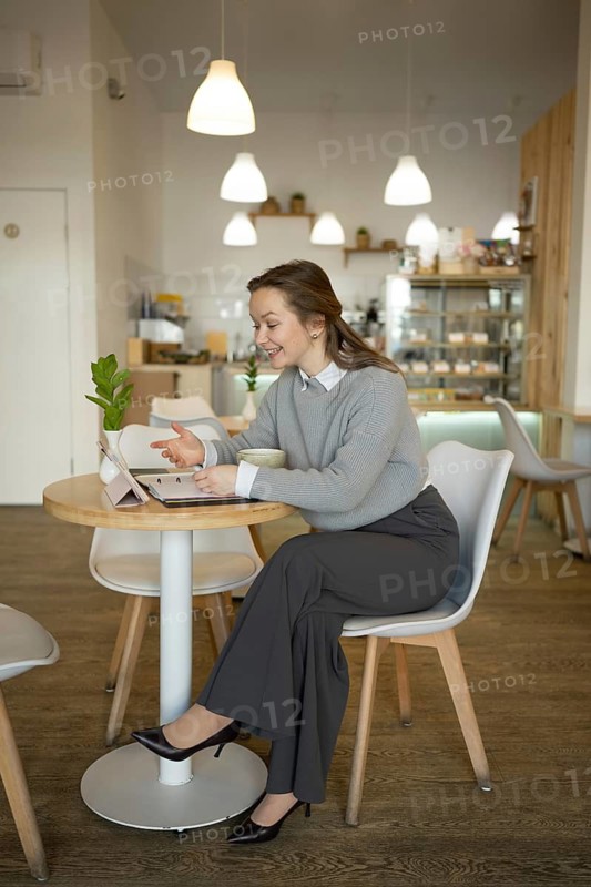 A woman in professional attire sits alone at a cafe table, engaging with a digital tablet. Surrounded by stylish decor and soft lighting, the scene conveys focus, comfort, and productivity