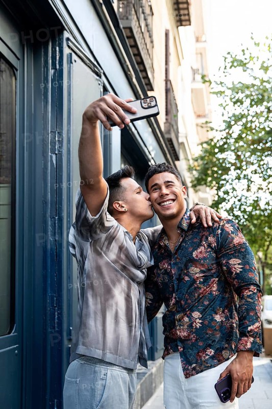 A cheerful latin gay couple takes a selfie while walking in the vibrant streets of Madrid, capturing a joyful moment with a tender kiss and bright smiles