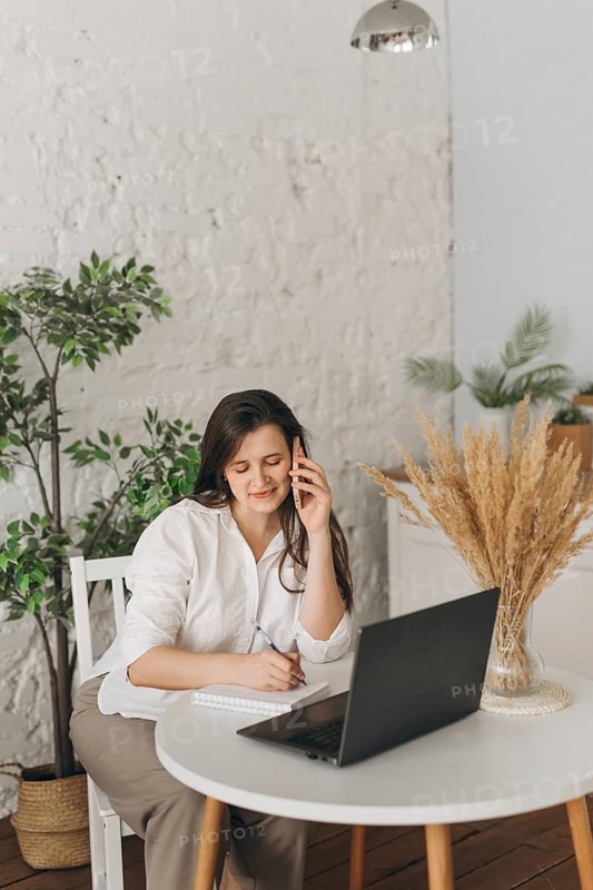 Nutritionist sitting at a white table, engaged in a call with a client. She's smiling, taking notes on a notepad, with a laptop open, in a cozy office setting with plants