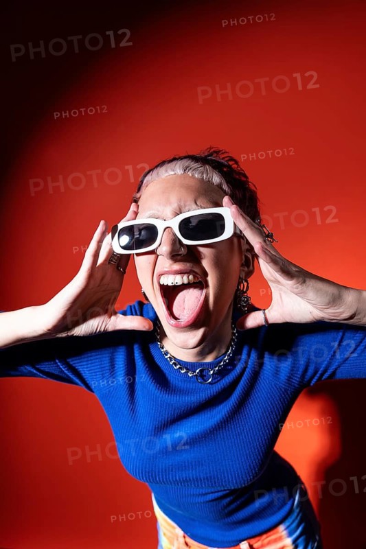 A trendy woman in colorful trousers, tooth gems and white sunglasses poses energetically against a bold red background, embodying modern fashion and vibrant self-expression