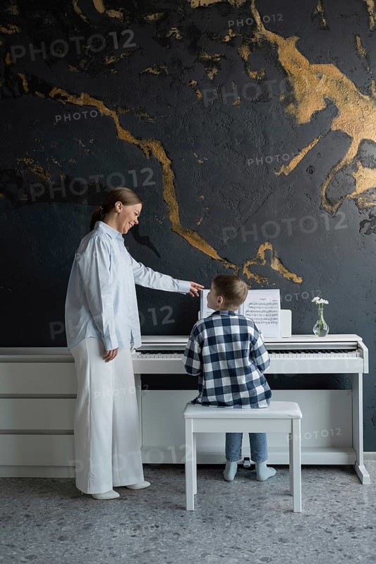 A young student receives piano instruction from a teacher in a cozy home setting. The child sits at a white piano, guided by the teacher pointing to sheet music on the stand