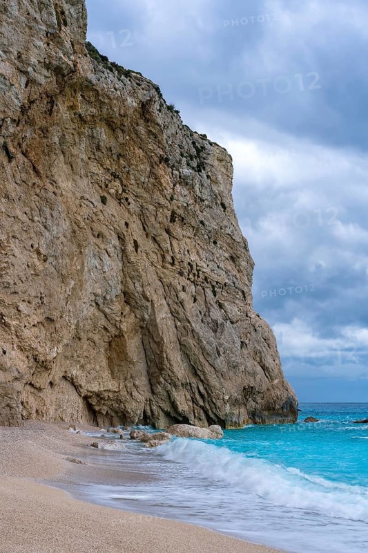 Porto Katsiki beach in Lefkada, Greece, features striking turquoise waters meeting rugged cliffs. Waves gently lap the sandy shore under a cloudy sky, creating a serene atmosphere