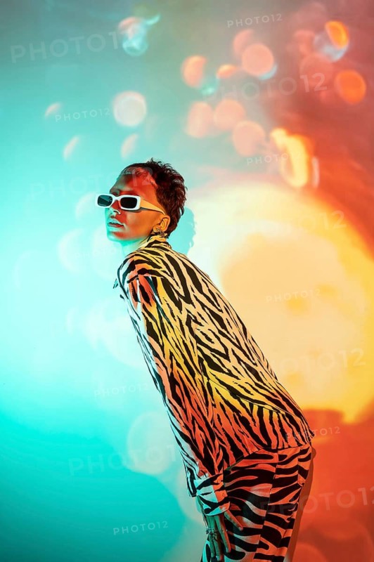 A young, trendy woman stands confidently in vibrant, colorful lighting. Her stylish sunglasses and bold, zebra-patterned outfit highlight a modern, fashionable aesthetic