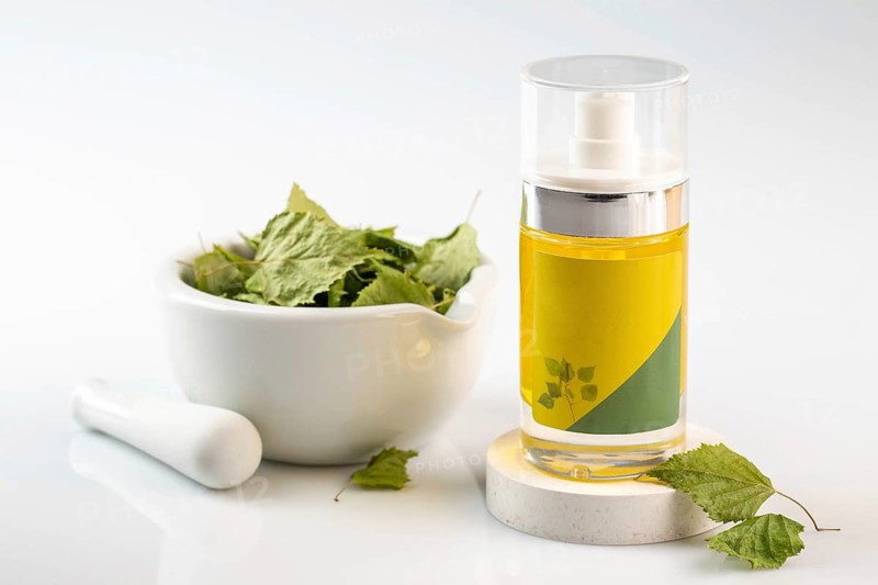 A clear bottle of birch oil stands beside a white mortar and pestle filled with green leaves, set against a clean white background, emphasizing natural skincare