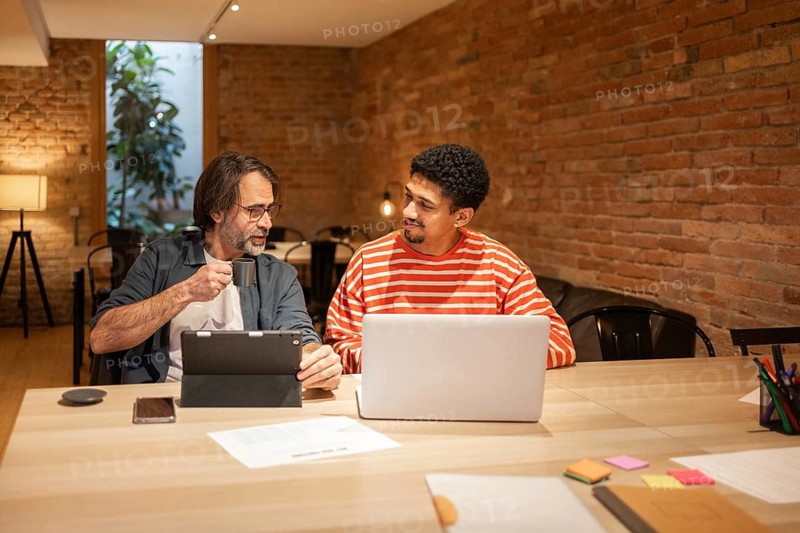 Two coworkers engage in discussion at a modern coworking space. One holds a tablet and coffee, the other uses a laptop. The setting features a brick wall and relaxed atmosphere