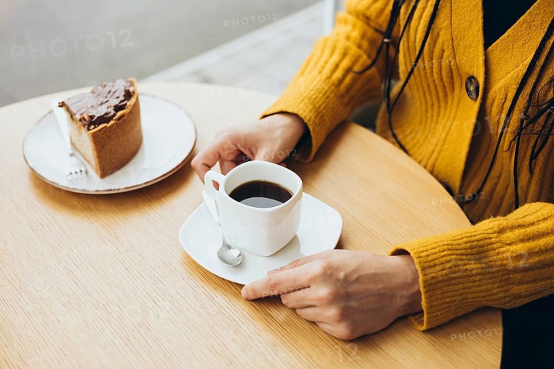 A person in a yellow sweater sits at a wooden table, savoring a cup of coffee alongside a slice of cake The scene evokes warmth and relaxation
