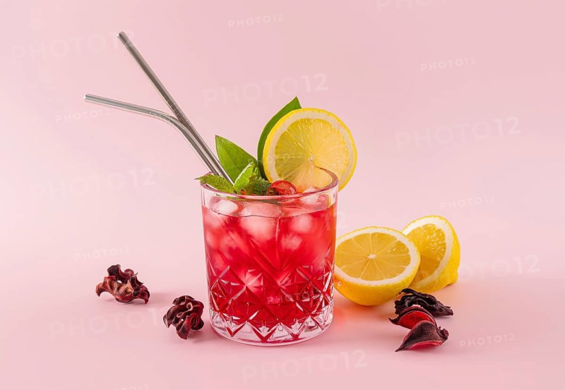 A vibrant red hibiscus drink in a crystal glass with ice cubes, garnished with lemon slices, mint leaves, and two metal straws, set against a soft pink background