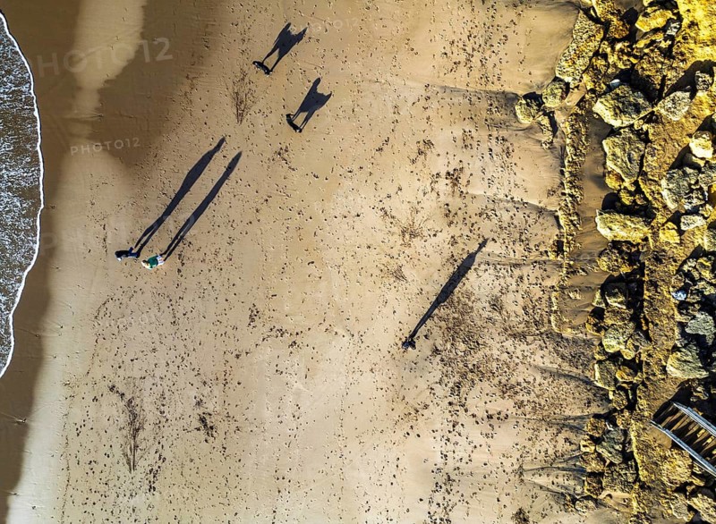 Aerial shot of a sandy beach with long shadows cast by people and dogs walking in Australia. Footprints trail behind them, and rocky formations line the shoreline