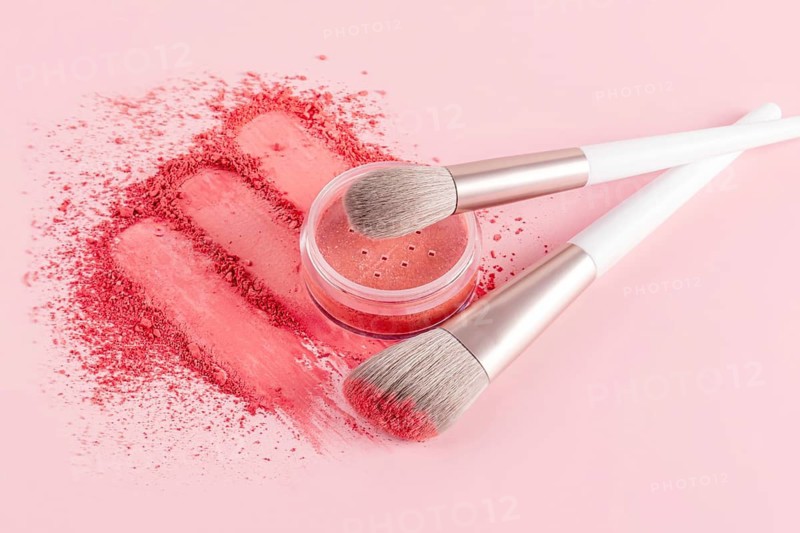 A vibrant makeup composition featuring a compact blush and two makeup brushes against a soft pink background. The blush is spread elegantly, highlighting its vivid color