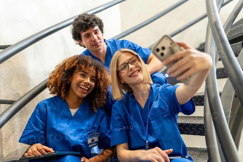 Three smiling medical students in blue scrubs are taking a cheerful selfie together on a metal staircase, representing friendship, teamwork, and diversity within healthcare education