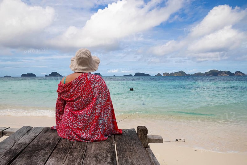 A woman in a sunhat and colorful sarong sits on a wooden platform, facing the azure waters and lush islands of Indonesia's Raja Ampat. The scene captures tranquility and adventure