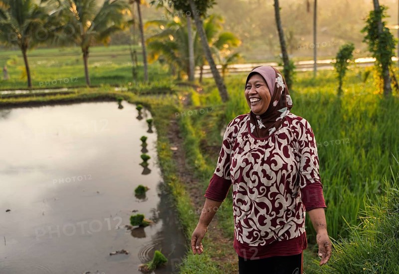 A smiling woman stands by a rice paddy in Indonesia, embraced by the warm glow of a sunset. Palm trees and vibrant rice fields create a serene, picturesque backdrop