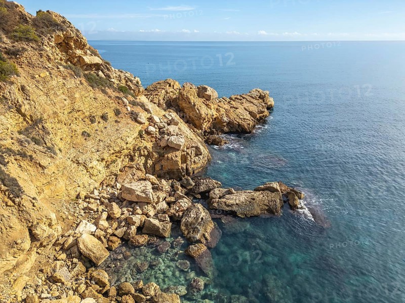 Stunning view of Costa Blanca's rocky coastline against a tranquil Mediterranean Sea. The rugged cliffs bathed in sunlight create a peaceful and picturesque landscape