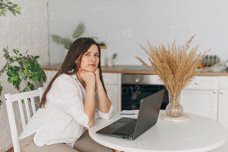 Nutritionist sits at a kitchen table, leaning on hands, with a laptop and notes in a modern home office setup. A vase of dried plants adds a natural touch to the scene