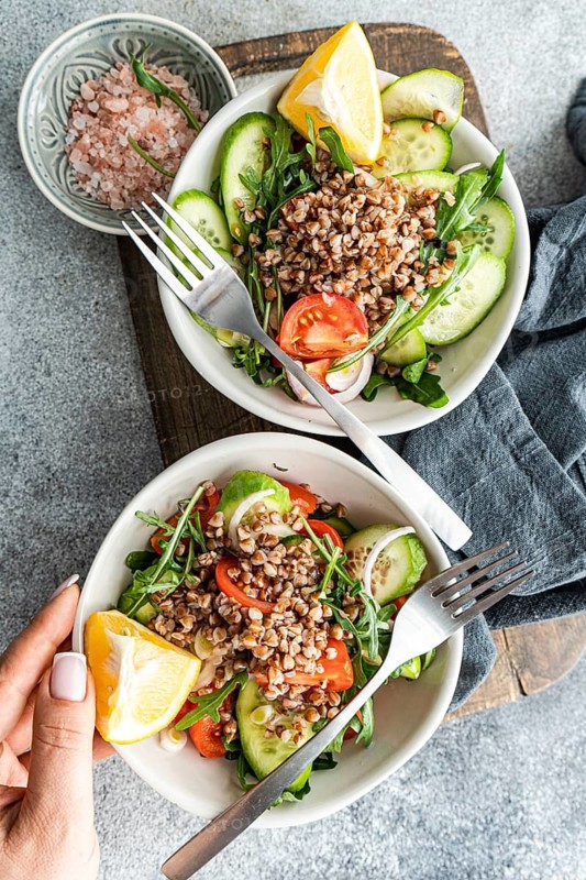 A vibrant, healthy lunch featuring fresh rocket, oregano, cherry tomatoes, cucumber, red onion, lemon, organic olive oil, Himalayan salt, and steamed buckwheat in a bowl