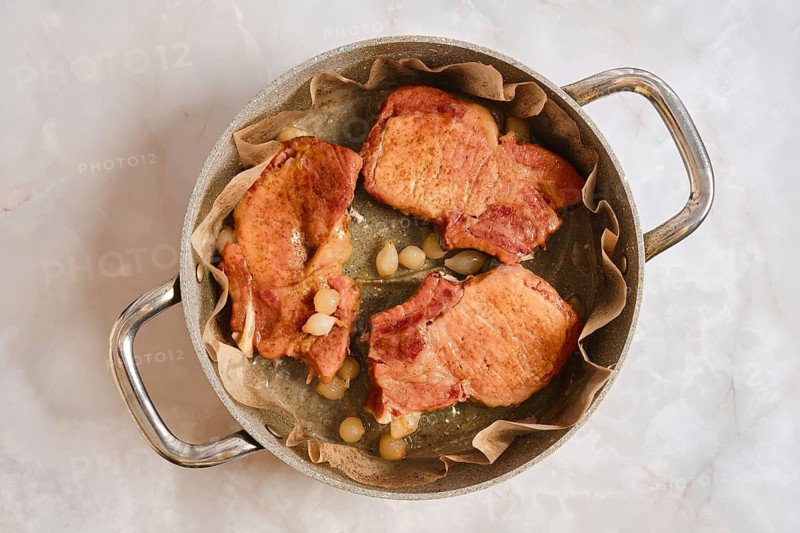 Roasted pork rib chops are placed in a pot lined with parchment paper. They are surrounded by sliced onions and are being prepared in a kitchen. The surface is a light marble
