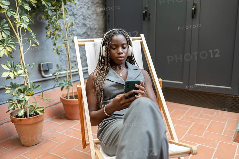 A young African woman with braids enjoys a summer day in a coworking space's outdoor area. She's seated in a lounge chair, using a smartphone and wearing headphones, surrounded by plants