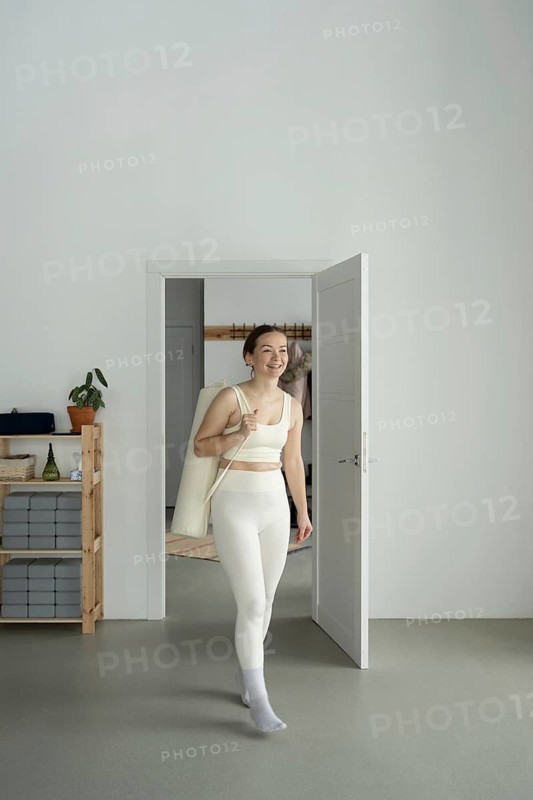 A woman in white active wear enters a yoga studio, carrying a mat bag over her shoulder. She exudes positivity and readiness for a yoga session, surrounded by minimalist decor