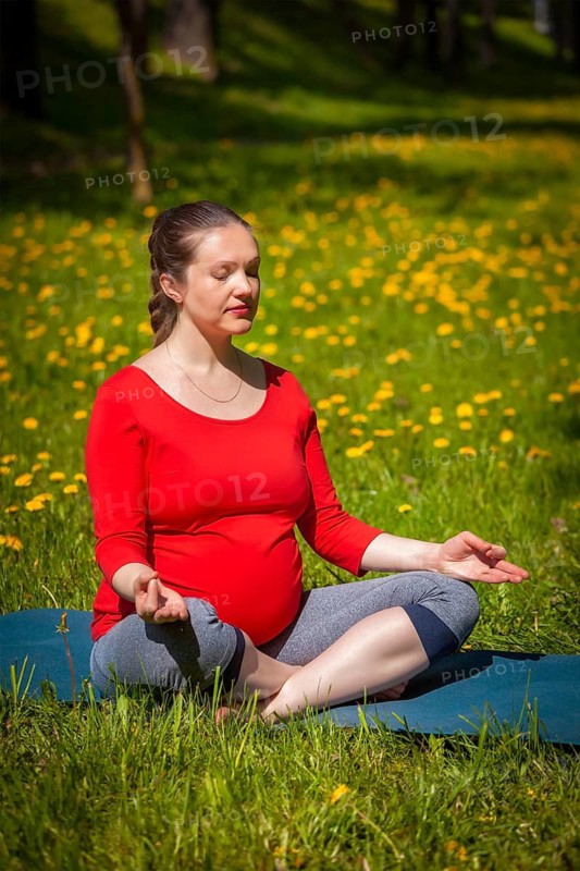 Pregnancy yoga exercise, pregnant woman meditating in yoga asana Sukhasana easy yoga pose with chin mudra outdoors on grass lawn with dandelions in summer