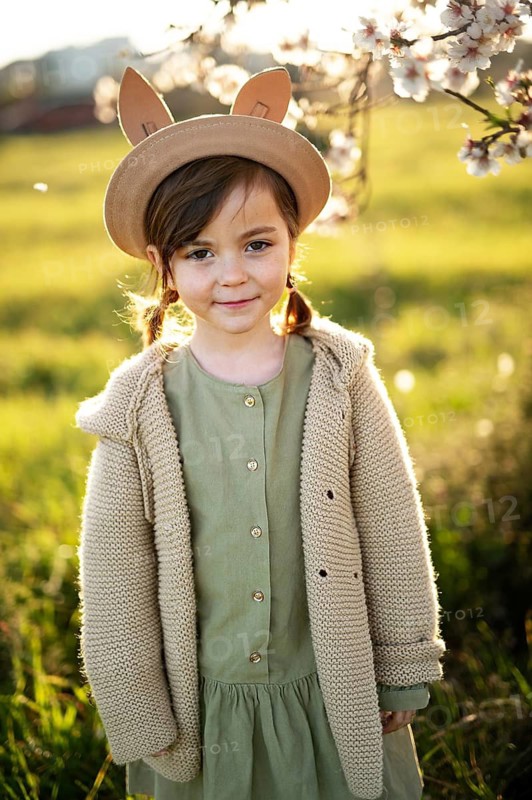 A young girl, dressed in a green dress and cozy sweater, stands in a field with almond blossoms in the background during springtime