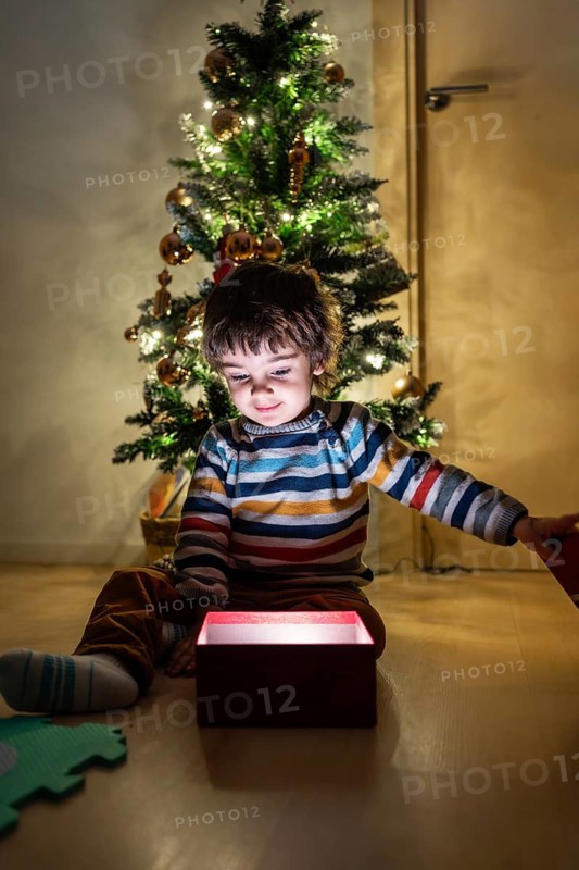 A young boy is sitting on the floor in front of a Christmas tree, opening a red box