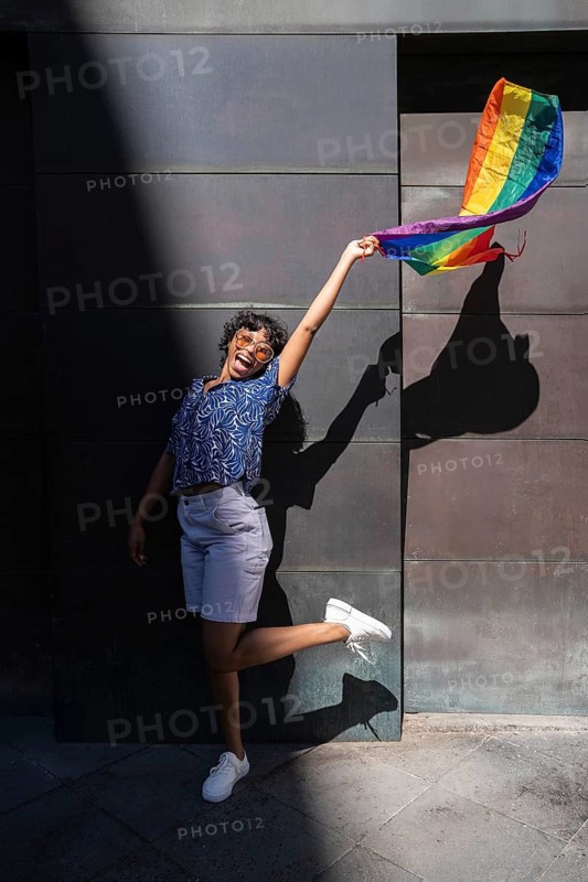 A joyful LGBTQ+ person wearing sunglasses holds a vibrant rainbow flag, casting a shadow against a dark urban wall, embodying freedom and pride on a sunny day