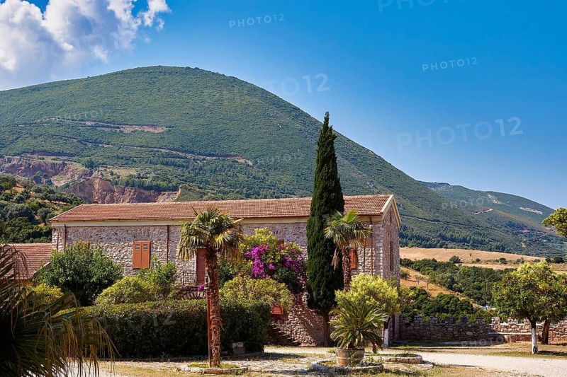 Rustic stone house in front of wooded hill under blue sky, surrounded by green vegetation, Achaia Clauss winery, Patras, Peloponnese, Greece