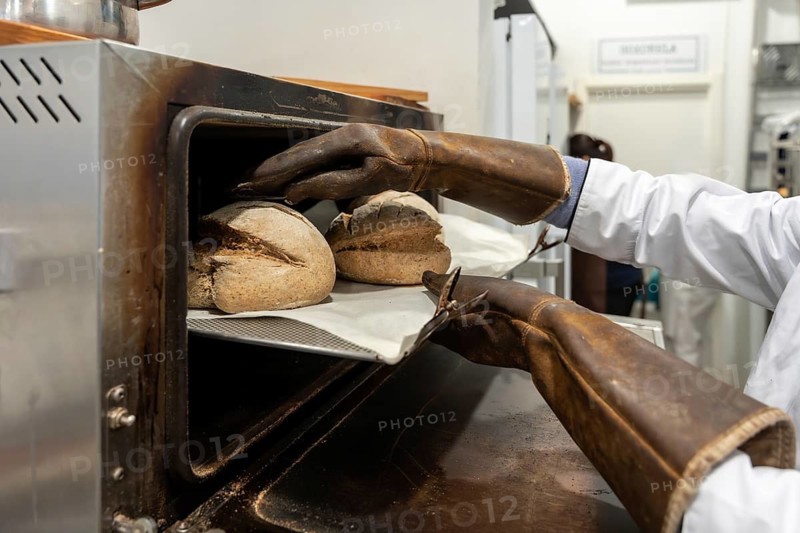 Baker wearing protective leather gloves removing freshly baked loaves of rustic bread from a professional convection oven, emphasizing traditional baking process and artisanal quality