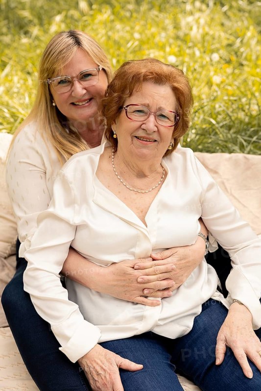 A joyful moment in the sun as a mother and daughter embrace, seated on a blanket with a backdrop of lush greenery. They share smiles and warmth, celebrating familial love