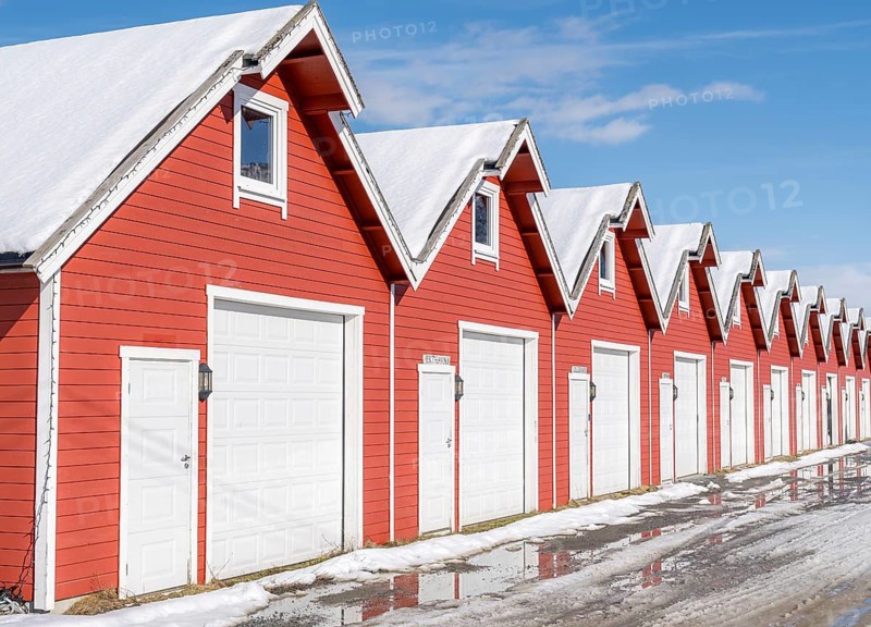 A row of red boathouses with snowy roofs reflects the traditional architecture of Botnhamn, Norway. The bright red color contrasts with the clear blue sky