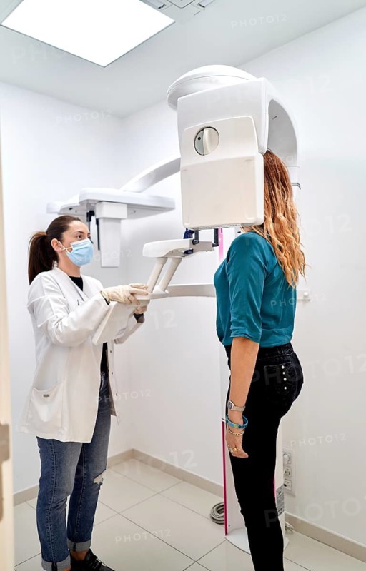 A female dentist in a white coat and mask assists a patient with a panoramic dental x-ray in a modern dental clinic, demonstrating advanced dental care technology