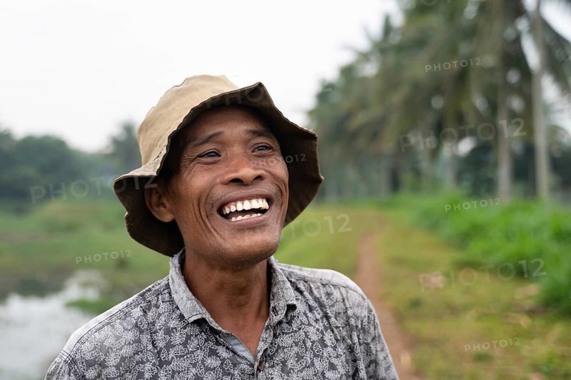 A joyous Indonesian farmer stands in a lush rice field, wearing a hat and patterned shirt. The serene landscape features greenery and palm trees, symbolizing rural life