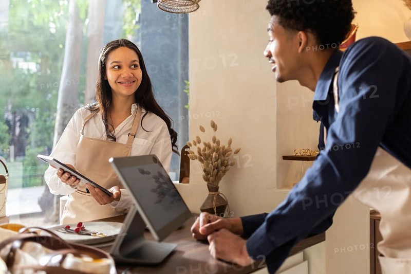 Two employees engage in a friendly conversation inside a cozy bag store. They are wearing aprons and using a digital tablet, surrounded by stylish bags and natural decor