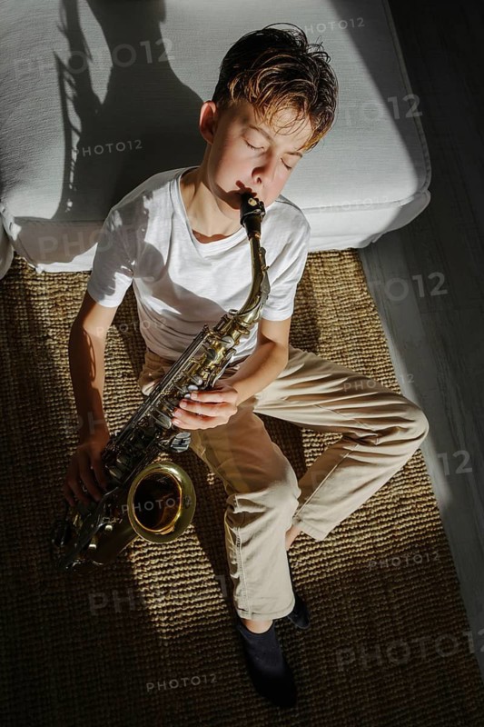 A young musician sits on a carpet, eyes closed, playing a saxophone. Sunlight creates a shadow, adding warmth and tranquility to the scene, evoking a sense of passion and focus